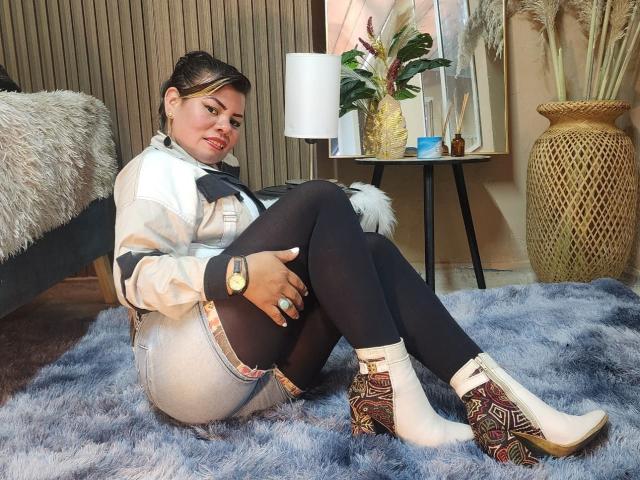 KiaraXMoon sits on a blue rug, legs crossed, wearing a white top, black leggings, and patterned boots. She has a sexy, relaxed pose with a slight smirk. Fucking hot.
