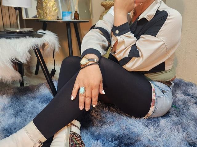 KiaraXMoon sitzt auf einem blauen, flauschigen Teppich, die Beine gekreuzt, und trägt ein schwarz-weißes Hemd, schwarze Leggings und weiße Socken. gestreifte Akzente. Ringe an ihren Fingern, Armbanduhr am linken Handgelenk.