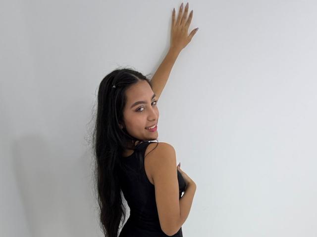 Emma Estreilla mit langen, schwarzen Haaren, die verführerisch lächelt, mit einem Arm angehoben vor einer weißen Wand, trägt ein schwarzes Top ohne Ärmel. von oben, Blick über die Schulter, hellbraune Haut, glänzliche Lippen, weiche Schatten.