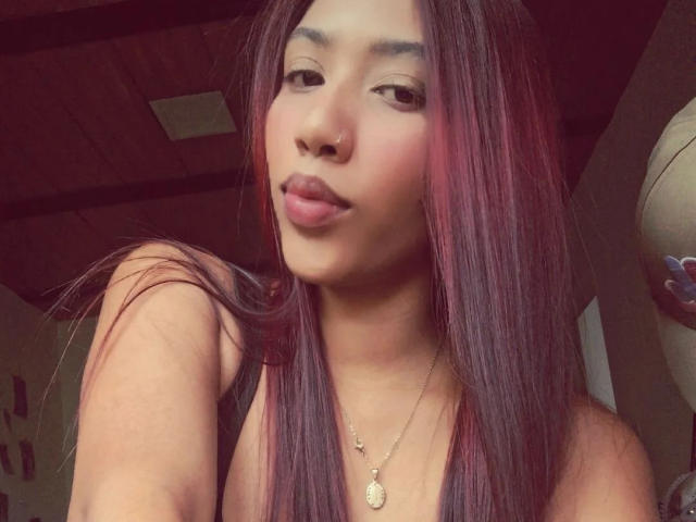 Close-up van GabiHott, een Aziatische vrouw met lang, steil, donkerroze haar, volle lippen en een gouden ketting. verleidelijk naar de camera kijkend. Ze heeft een neusring en een gladde huid..