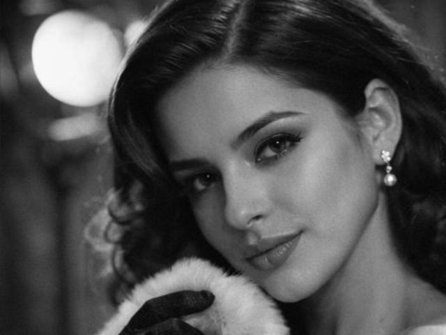 Black and white photograph of IsabellaJames, close-up, holding a fur-trimmed garment near her face. She has wavy hair, dark makeup, and pearl earrings. She's looking directly at the camera with a slight smirk.