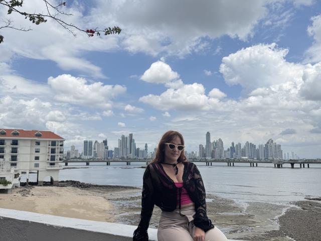 JaneRoa, com cabelo castanho e óculos de sol, vestindo uma jaqueta preta e uma blusa rosa, sentada em uma calçada perto de um cais, na cidade. silhueta e céu nublado ao fundo. Muito sexy..