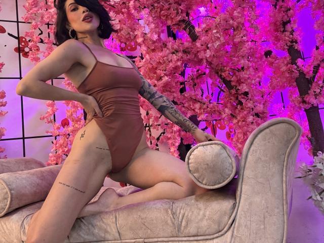 HannaBorbon está arrodillada sobre una chaise lounge color beige, vistiendo un traje de baño entero marrón ajustado. Ella tiene el pelo oscuro, labios rojos y un tatuaje en su brazo derecho. Fondo de flores de cerezo rosadas que brillan con un tono púrpura..