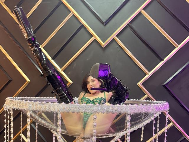 HannaBorbon sits in a glittering, crystal-encrusted chair, legs spread wide, wearing green lingerie and a black hat, holding a large black gun, against a geometric, black-and-gold wall. Fucking hot.