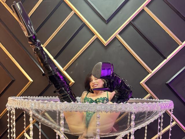 HannaBorbon, femme séduisante, jambes écartées, lingerie verte, bottes hautes noires, tenant un pistolet noir, assise sur un cristal. lustre, mur en chevron noir et or, reflet violet dans le miroir, pose séduisante.