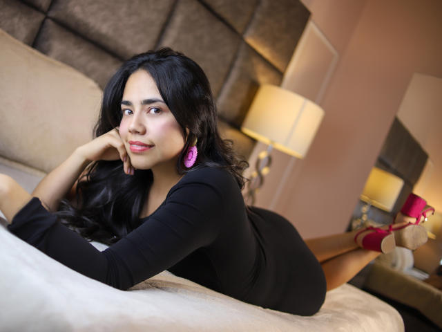 Sophie Weber ligt op een bed, met haar benen gebogen, en draagt een strak zwart jurkje, roze oorbellen en rode hakken. Ze leunt haar hoofd op haar hand en kijkt verleidelijk naar de camera..