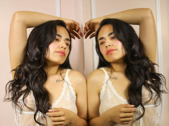 Fotografía de Sophie Weber con cabello negro largo y ondulado, vistiendo una blusa blanca de encaje, con los brazos levantados, reflejo en un espejo. Luz suave, expresión segura, tatuaje discreto en el pecho. Increíblemente sexy, pose reflejada..