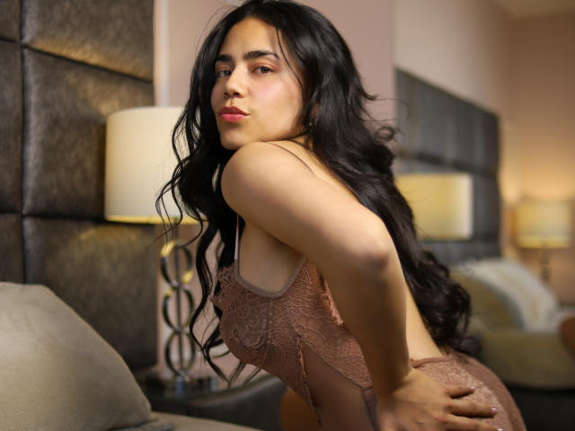 SophieWeber, a sexy Latina woman with long black hair, leans forward, hands on her ass, wearing a glittery, pink lingerie dress. She has a seductive look, with a visible cock in the background.