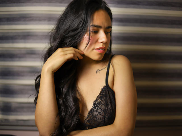 Fotografia di Sophie Weber, una sexy donna latina con lunghi capelli neri, che indossa un reggiseno in pizzo nero e guarda in basso. un sorriso appena accennato.
la sua mano destra tocca il mento. Sfondo a righe sfocate..