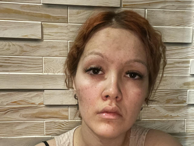 Nahaufnahme von AgathaMeler, einer blassen Frau mit roten Haaren, Sommersprossen und schwarzem Eyeliner, vor einer Holzauskleidung. Wand. Sie blickt direkt in die Kamera, die Lippen sind leicht geöffnet, und sie wirkt gelangweilt. Ihre Schulter ist sichtbar, sie trägt ein helles Oberteil..