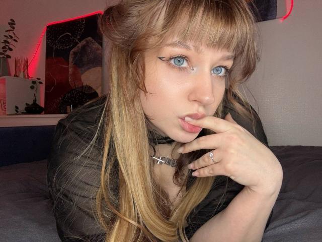 TheressaBuzzo has bright blue eyes and light brown hair with bangs, biting her finger seductively. She wears a black top, a cross necklace, and a ring, with a sexy expression. Red neon lights in the background.