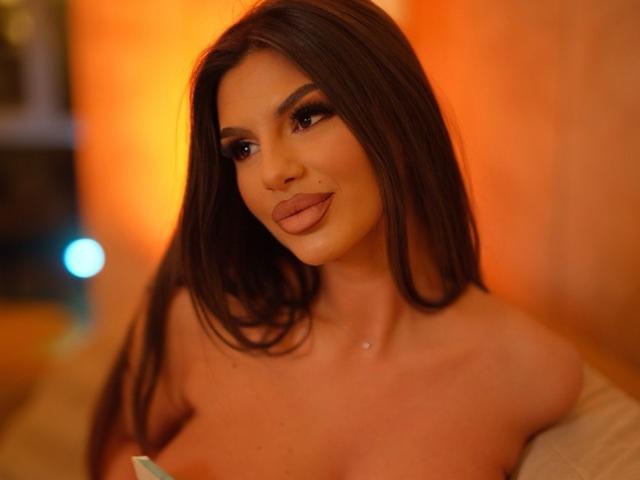 Photograph of JessyKiss, topless with long brown hair, full lips, and dark eyes, illuminated by warm orange light. JessyKiss has a slight smile, with a subtle nose piercing. Background is blurred.