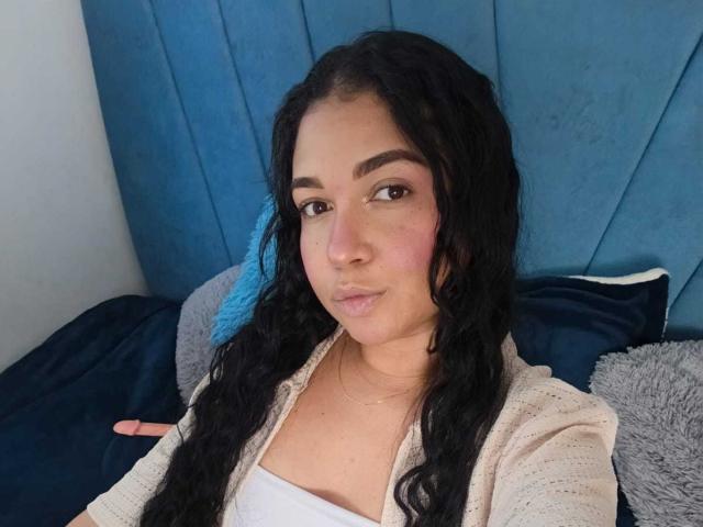 Alice Smity, com cabelos pretos longos e ondulados, olhos castanhos e bochechas rosadas, vestindo uma blusa branca e um cardigã bege, sentada. contra um cabeceira azul, expressão sensual, lábios ligeiramente entreabertos, olhar sugestivo.