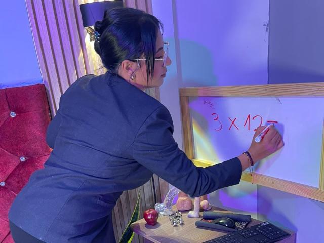 DiannaRosse, avec ses cheveux sombres attachés en chignon, portant un blazer noir, écrivant "3 x 4 = 12" sur un tableau blanc avec un stylo rouge. Chaise rouge, bureau avec une pomme, lampe. Tension sexuelle.