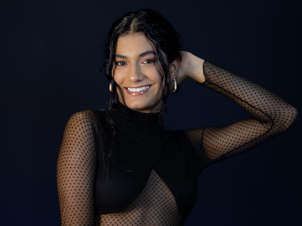 Fotografía de HannaCollyns, sonriendo con dientes blancos brillantes, cabello oscuro, vistiendo una blusa negra de malla transparente, con una mano detrás. cabeza, sexy, fondo oscuro, iluminación intensa, pose segura, atractivo/a..