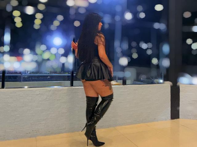 ArtemisaStone è in piedi su un balcone di notte, di spalle, indossando una gonna di pelle nera e stivali neri alti fino alla coscia. Capelli lunghi e neri.
le luci della città si confondono sullo sfondo. Incredibilmente sexy..