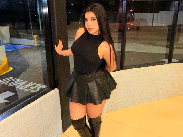 ArtemisaStone, con el pelo largo y negro, vestida con una camiseta negra sin mangas y una falda de cuero negra corta, botas hasta la rodilla, de pie frente a una ventana de vidrio, con la mano sobre el vidrio, fondo de la ciudad por la noche..