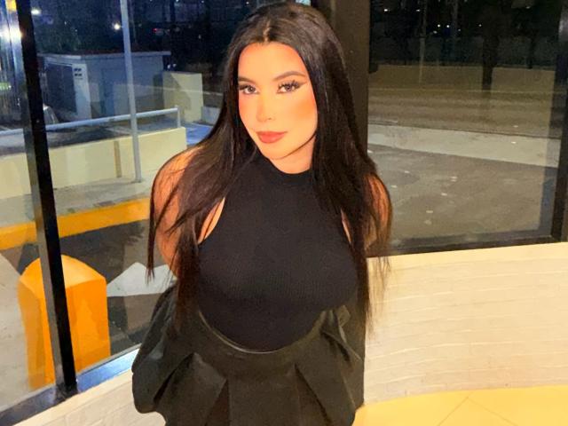 ArtemisaStone, con el pelo largo y negro, vestida con una camiseta negra sin mangas, está de pie frente a una ventana de vidrio por la noche. Su expresión es segura, con una sutil tensión sexual. Elementos de fondo de color naranja brillante..