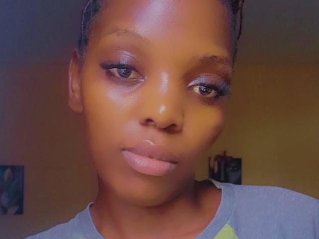 Close-up of Gladwell, an attractive Black woman with smooth, dark brown skin and full lips, wearing a light blue shirt. Her eyes have smoky eyeliner, and she's looking directly at the camera with a subtle, confident expression.