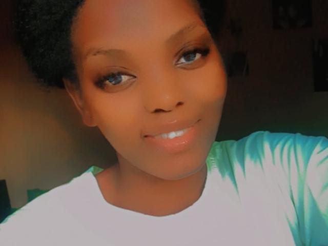 Close-up selfie of Gladwell with dark brown skin, blue eyes, and glossy lips. Gladwell looks directly at the camera with a subtle smile, wearing a white shirt. Glowing light highlights face.