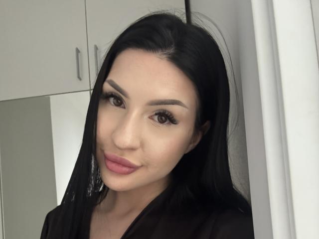 AuraXo, avec de longs cheveux noirs, des yeux marrons et des lèvres roses, regarde directement l'appareil photo avec un léger sourire. Elle porte un haut noir et se tient devant un meuble blanc. Extrêmement sexy..