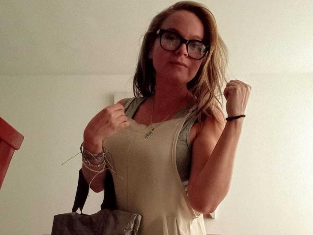 Fotografia de Pamela Colin com cabelo loiro na altura dos ombros, usando óculos e uma camiseta bege, segurando uma bolsa cinza. pulseiras em seu pulso, olhando diretamente para a câmera com um sorriso discreto..