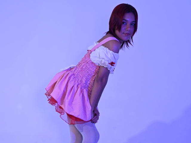 NikkyDoll beugt sich vor und trägt ein verspieltes Lächeln, gekleidet in einem rosa Karomusterkleid mit weißen Ärmeln. Ihre kurzen, braunen Haare und das blaue Licht erzeugen eine sexy, provokante Atmosphäre. Total süß..