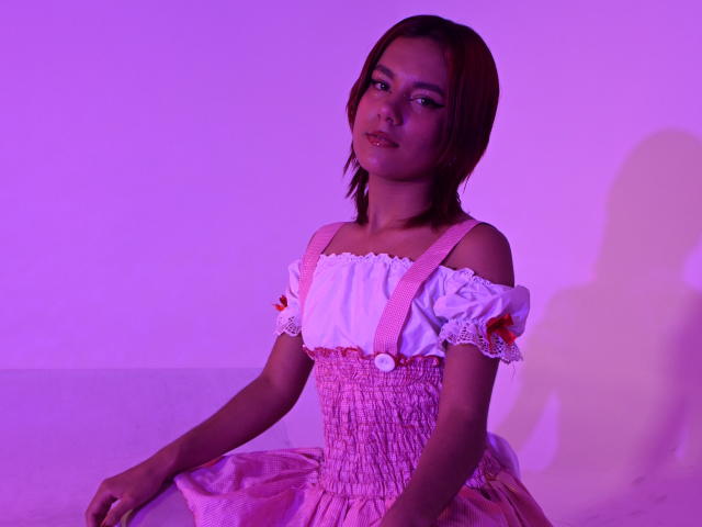 NikkyDoll sits with a relaxed pose, wearing a pink off-shoulder dress with white puffed sleeves, bathed in purple and pink lighting. She has wet, shoulder-length hair and a subtle smile.