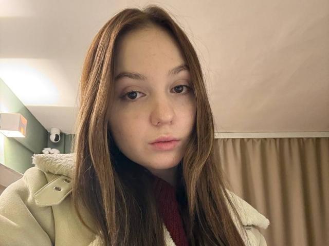 Close-up van BaoRoebke met lang bruin haar, gekleed in een beige jas, die recht in de camera kijkt met een neutrale uitdrukking. uitdrukking.
zachte, warme verlichting binnen, een beige gordijn op de achtergrond, een lichte seksuele spanning..