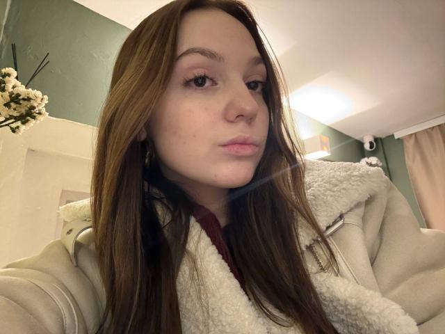 Autofoto de primer plano de BaoRoebke, una joven con cabello largo y castaño, que lleva un abrigo beige con un cuello blanco de piel sintética. Mirando directamente a la cámara con una expresión neutra. Luz suave en el fondo.