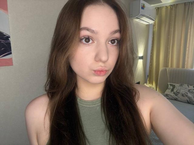 Photograph of BaoRoebke with long brown hair, pale skin, and pouty lips, wearing a green sleeveless top. Background includes a lit room with curtains and a sofa. Fucking cute, sexy vibe.