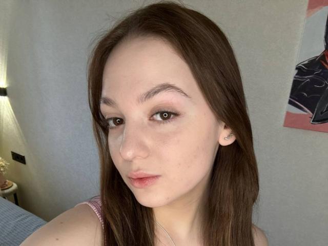 Close-up of BaoRoebke with fair skin, brown hair, and brown eyes, looking directly at the camera with a neutral expression. Wearing a pink spaghetti strap top. Soft natural light.