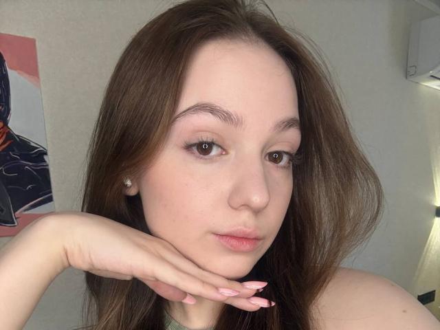 Close-up of BaoRoebke, a fair-skinned woman with long brown hair, resting her chin on her hand, showing off pink nail polish. She has small earrings, brown eyes, and a neutral expression. Fucking sexy.