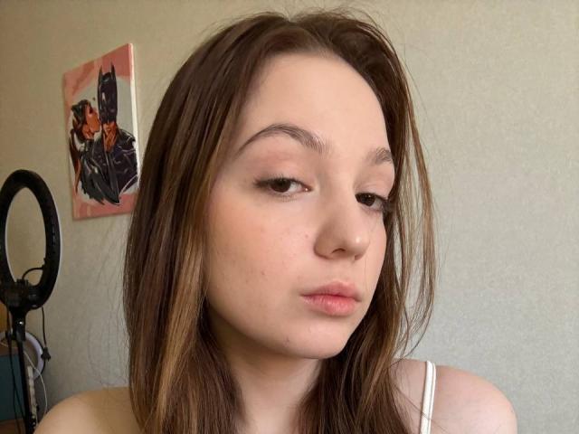 Primer plano de BaoRoebke, con piel clara, cabello castaño y expresión neutra. BaoRoebke tiene labios carnosos, una nariz pequeña y un maquillaje ligero. El fondo tiene una luz de anillo y una obra de arte colorida en la pared. Los ojos de BaoRoebke están medio cerrados, lo que le da un aspecto seductor..