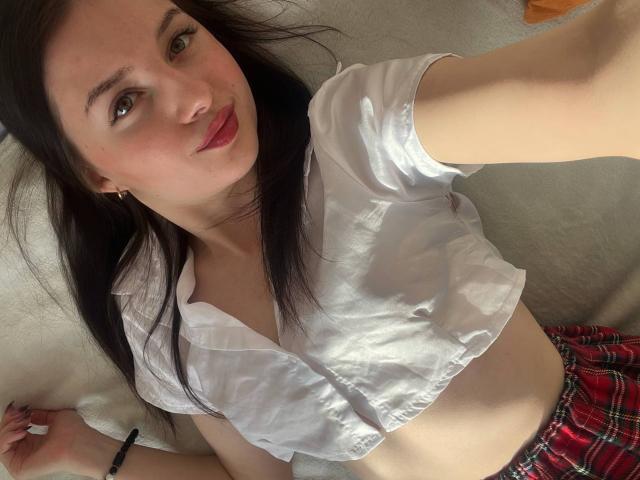 DollieSkweres lag auf einem Teppich und machte ein Selfie. Sie hat langes, schwarzes Haar, rote Lippen und trägt ein weißes, halbtransparentes Crop-Top mit einem roten karierten Rock. Das Sonnenlicht betont ihre helle Haut..