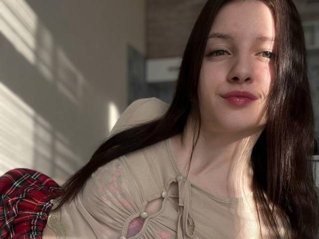 Close-up van DollieSkweres, een jonge vrouw met lang zwart haar, lichte huid en rode lippen, die een beige top met roze accenten draagt. bloemenborduurwerk en een rode geruite rok, zonlicht op haar gezicht.