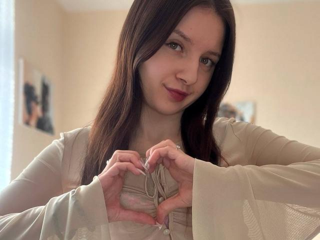 DollieSkweres, with long brown hair and fair skin, makes a heart shape with her hands in a beige top. She has a subtle smile and light makeup. Background is blurred.