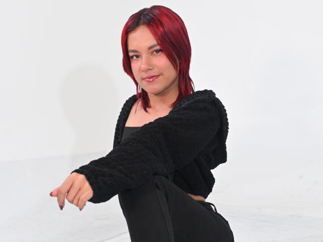 Digitale kunst van NikkyDoll met rood haar, die een zwarte, pluizige top draagt en vooruit wijst met een lichte grijns. Witte achtergrond, zwarte rok, gemiddelde lichaamsbouw, lichte huid, nonchalante pose..