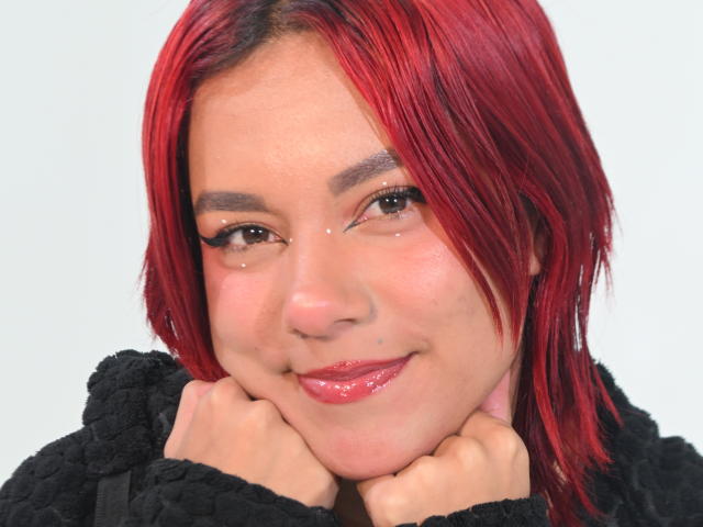 Close-up van NikkyDoll met levendig rood haar, lachend met haar handen onder haar kin. Ze heeft een lichte huid, donkere wenkbrauwen en glanzende rode lippen. Ze draagt een zwarte, pluizige top. Heel aantrekkelijk..
