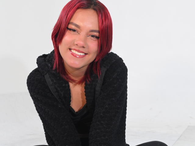 Photographie de NikkyDoll, souriante, avec des cheveux roux, penchée en avant, portant un sweat à capuche noir. Fond blanc éclatant, tension sexuelle, légère rougeur, contact visuel direct, décontracté, cheveux légèrement en désordre..