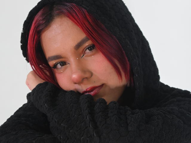 Primo piano di NikkyDoll con capelli rossi e neri, che indossa una felpa nera e pelosa. È china in avanti, con la mano sotto il mento, le labbra rosse leggermente dischiuse, e i suoi occhi marroni guardano seducentemente la fotocamera..