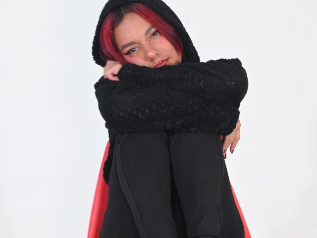 NikkyDoll, una giovane donna con i capelli rossi, si rannicchia in una giacca nera con cappuccio e pantaloni neri, con le gambe strette al petto, indossa un sfondo, sguardo intenso, sottile tensione sessuale, mantello rosso.