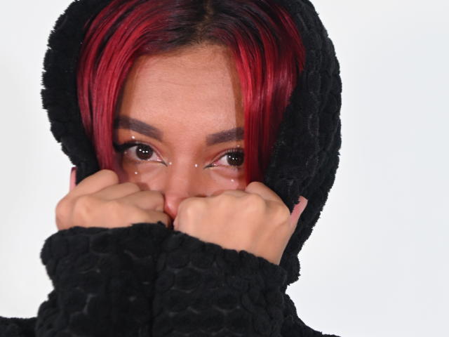 Close-up photo of NikkyDoll with red and black hair, wearing a black fuzzy hood. NikkyDoll's hands cover her mouth, eyes wide and seductive, glistening with sexual tension. White background.