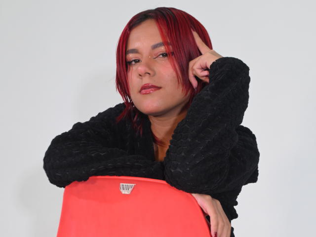 NikkyDoll met rood haar, een zwarte, pluizige jas en een feloranje top, die naar haar slaap wijst met een verleidelijke uitdrukking. witte achtergrond, digitale kunst, nonchalante pose, sexy, zelfverzekerde uitstraling.