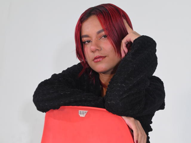 NikkyDoll met rood haar, die een zwarte, pluizige jas draagt en een felrode object vasthoudt. Ze leunt voorover, haar hand is dicht bij haar gezicht, en ze straalt seksuele spanning uit met een verleidelijke blik..