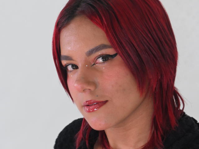 Close-up van NikkyDoll met rood haar, dikke zwarte eyeliner en glanzende rode lippen. Ze heeft een lichte glimlach, draagt een zwarte top, en haar poriën zijn zichtbaar op haar gezicht. Heel aantrekkelijk.