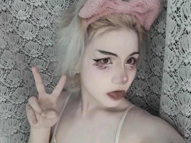 Gros plan de KatlynRenteria, avec une peau pâle, des cheveux blonds platine et un nœud rose. Elle fait un signe de la main en signe de paix, avec un trait d'eye-liner audacieux et un rouge à lèvres foncé, avec un fond en dentelle. Tellement mignon..