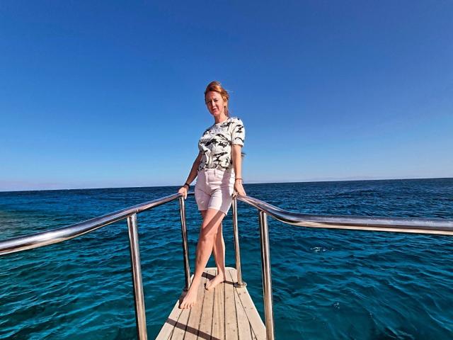 Tamariza stands confidently on a wooden pier, gripping metal railings, wearing a white graphic tee and shorts, blue ocean stretching behind her, bright clear sky above, sunlight on her skin.