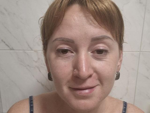 Primer plano de Tamariza, una mujer de piel clara con cabello corto castaño, un pequeño piercing en la nariz y aretes negros. Ella está sonriendo ligeramente, y lleva una camiseta sin mangas de color azul marino. Fondo de mármol blanco. Muy atractivo/a..