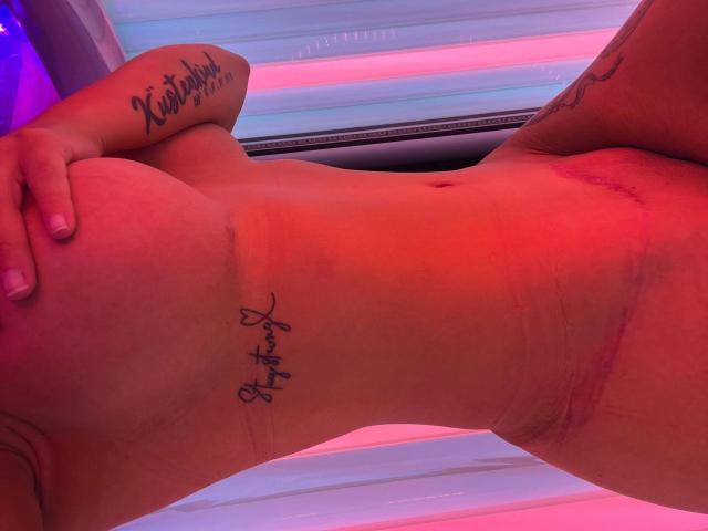 Nahaufnahme eines Fotos von SarasHotDiamond-hots nacktem Oberkörper, die in einem Solarium liegt, umgeben von rosa und blauen Lichtern. Sie hat Tattoos an ihrem Arm und an der Seite. Ihre Hand ruht auf ihrem Bauch. Ihre Haut glänzt im Licht..